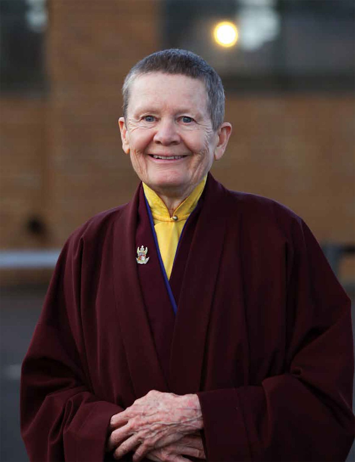 Pema Chodron standing and smiling. Her hands are crossed and she is wearing maroon robes and a yellow shirt underneath. She has short hair and bright eyes.