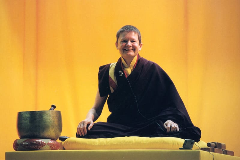 Pema Chodron smiling. She is sitting cross-legged on a pillow and is wearing maroon robes with a mustard yellow shirt underneath. She is against a yellow background.