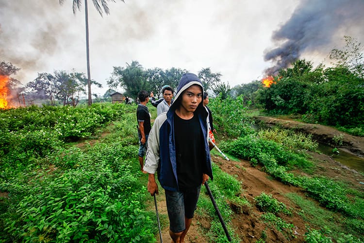 A young person walking through a grassy field. There are three more young people behind them and there are fires in the background.
