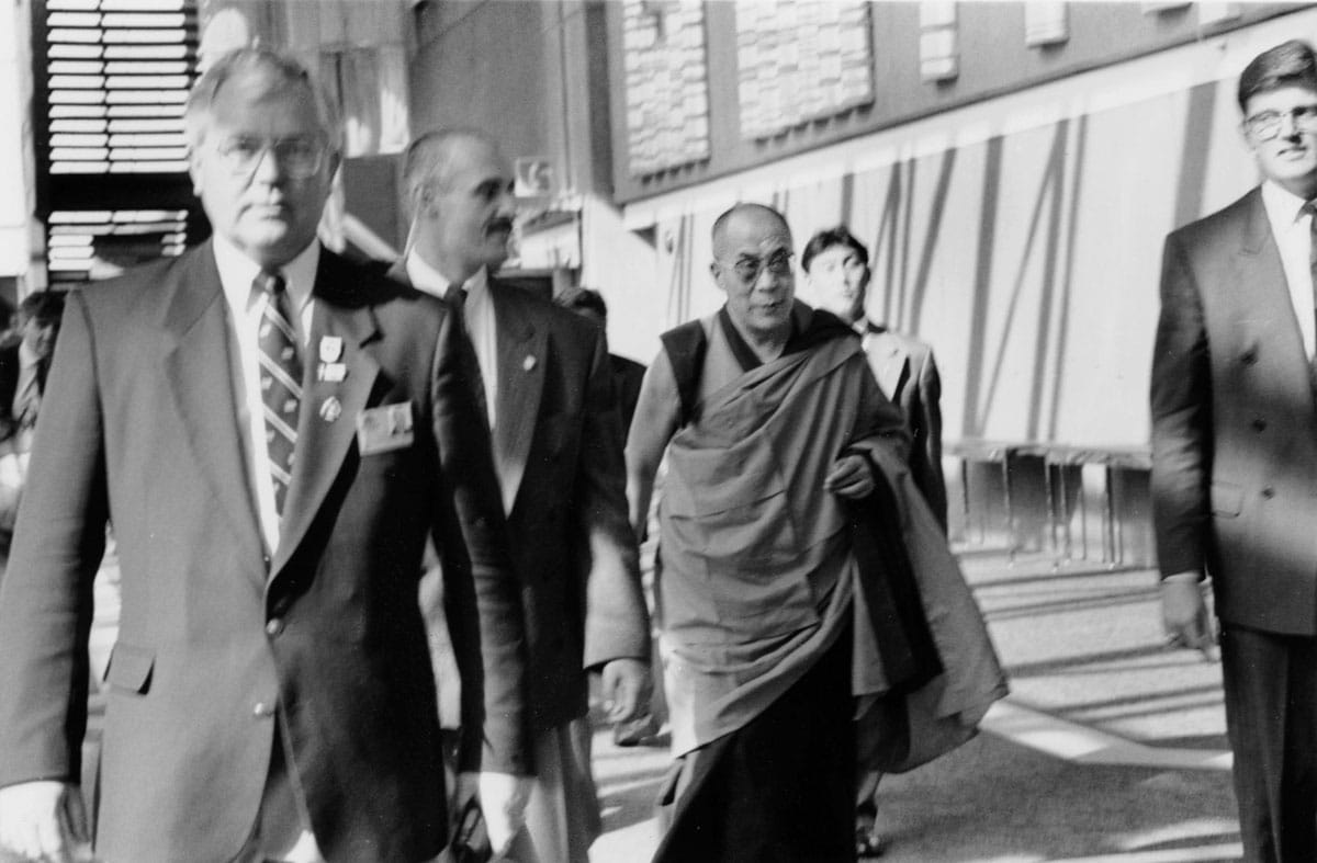 A black and white photo of the Dalai Lama walking through the airport. He is dressed in robes and is surrounded by people in suits.