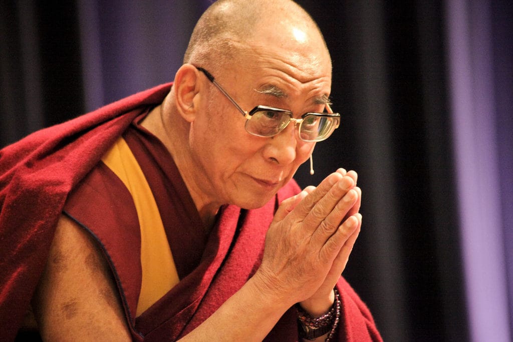 The Dalai Lama bowing. He is wearing red robes and has his hands placed together.