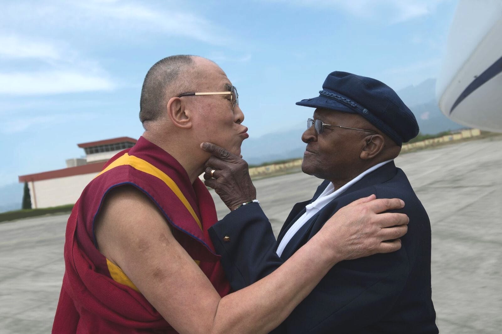 The Dalai Lama and Archbishop Desmond Tutu. The Dalai Lama is wearing marroon robes with a mustard yellow sash. Desmond Tutu wears a suit and a black hat.