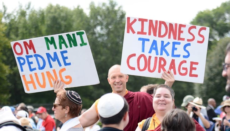 A Buddhist monk stands in a crowd holding one sign that says "Om Mani Padme Hung" and another that says "Kindness takes courage."