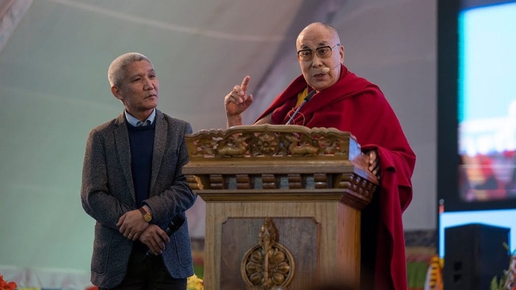 Dalai Lama speaking at lectern with Thupten Jinpa.