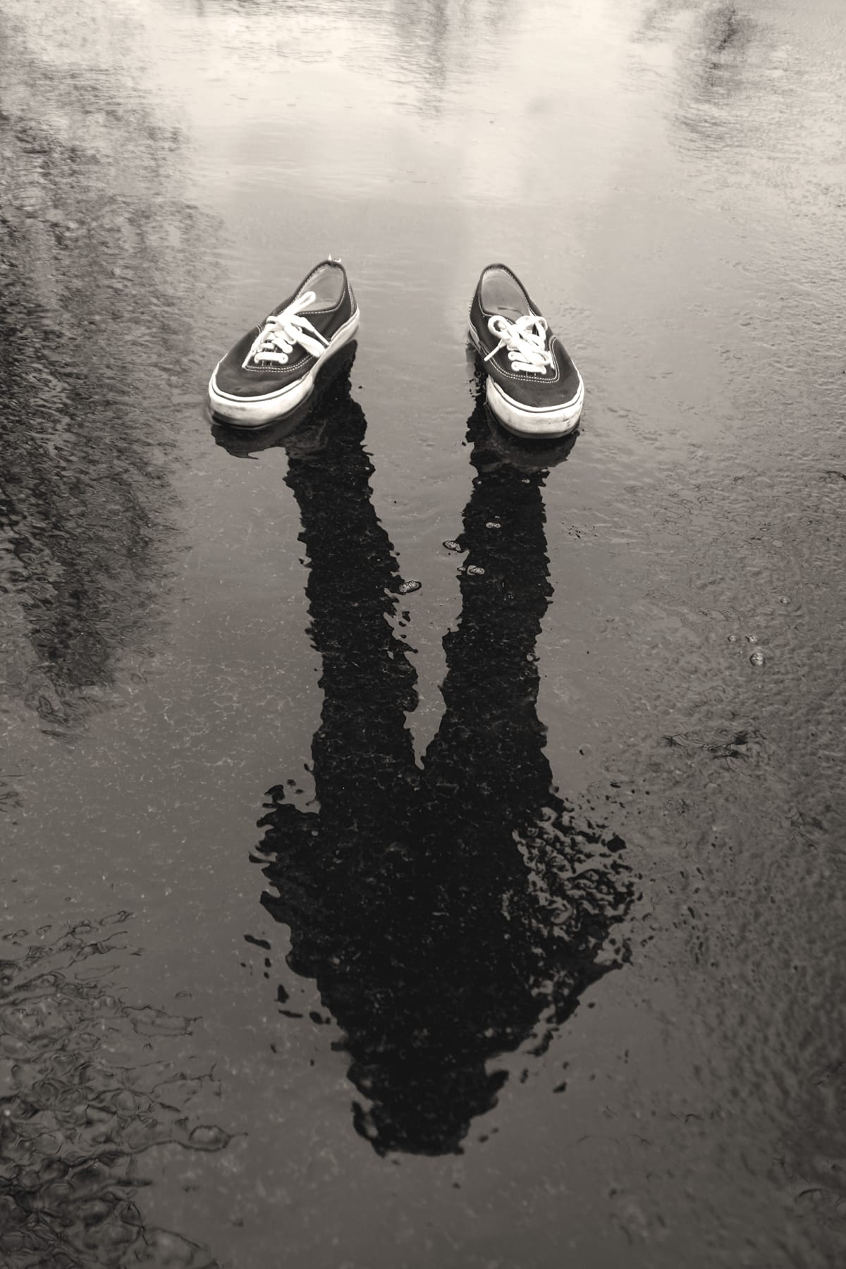 A black-and-white photo of a man's reflection on wet pavement with a pair of shoes on top of the reflection.