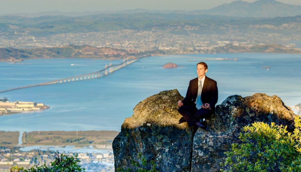 Man in a business suit sitting on a hilltop, meditating, with city skyline in background.