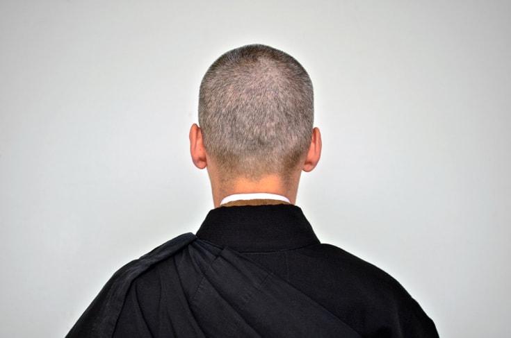 A photo of a monk taken from behind. Only the back of the head and the black robe is visible. Photo is taken against a grey wall.
