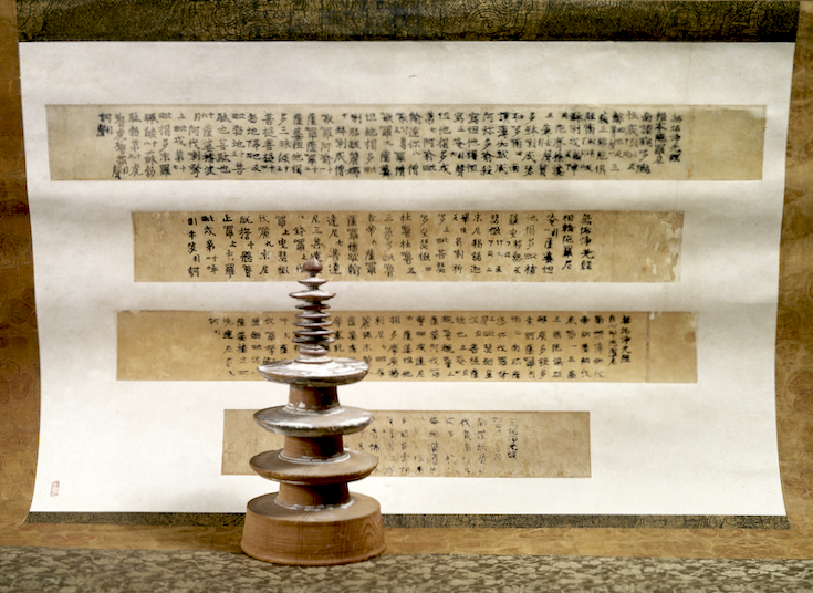 A small stupa stands in front of four pages of printed scripture.