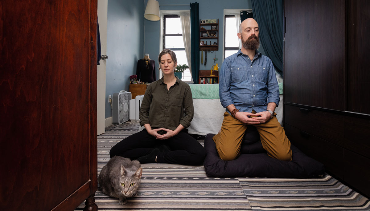 A couple meditating next to a cat. They are in a bedroom and sitting on a striped rug.