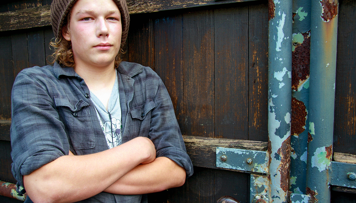 A young boy with a plaid shirt and hat stands with his arms crossed. 