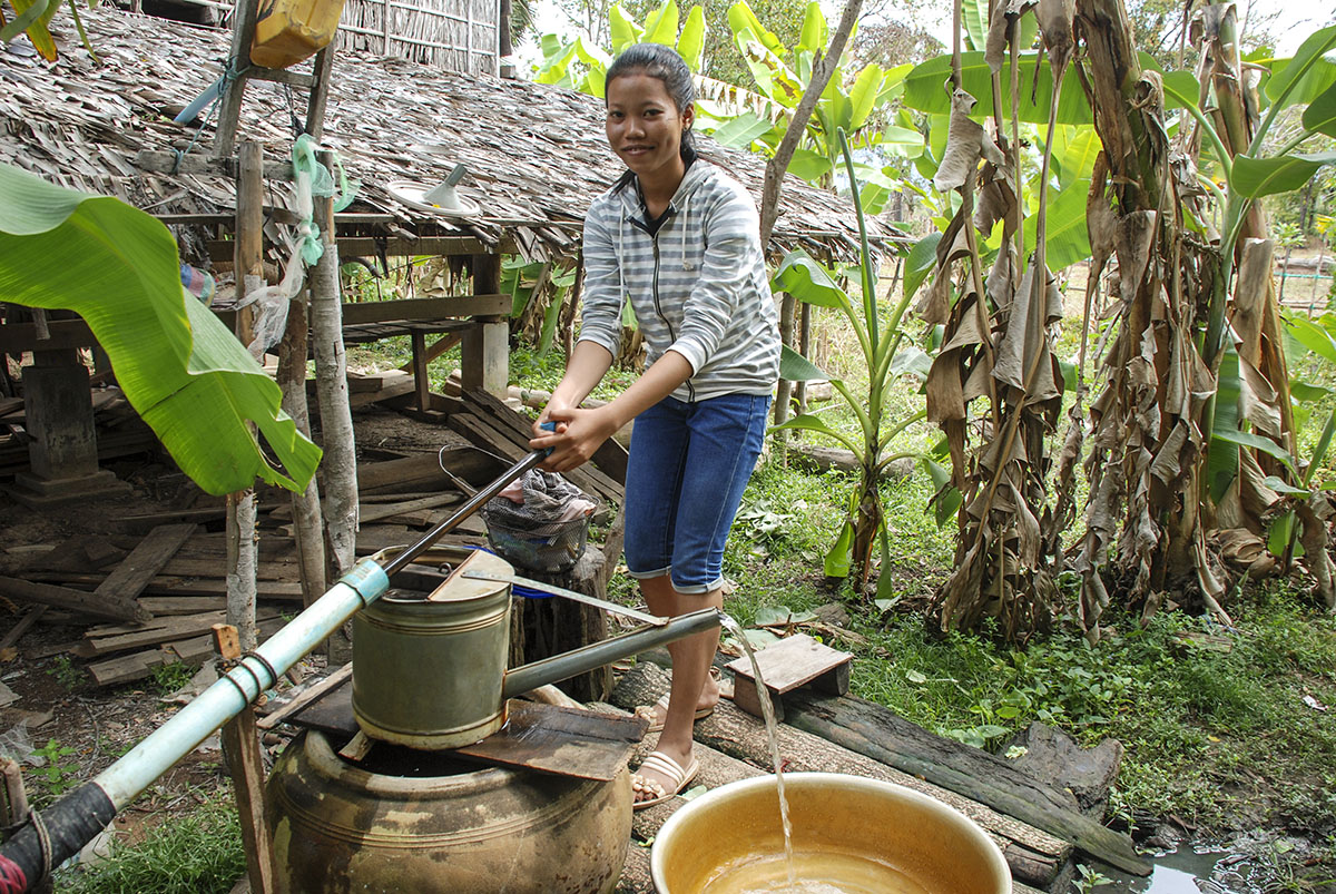 A woman wearing a striped hoodie and long jean shorts holds a water pump. There are lots of plants behind her.