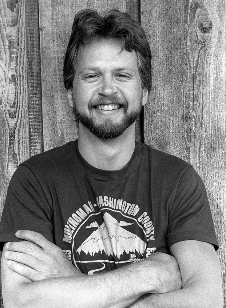 A black and white photo of a man wearing a tshirt. He is smiling with arms crossed against a wooden wall.