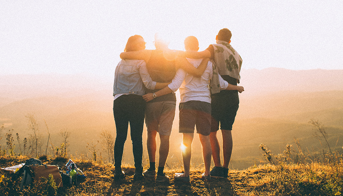 A photo of four people with their arms around each other looking off of a cliff. The photo is taken from behind.