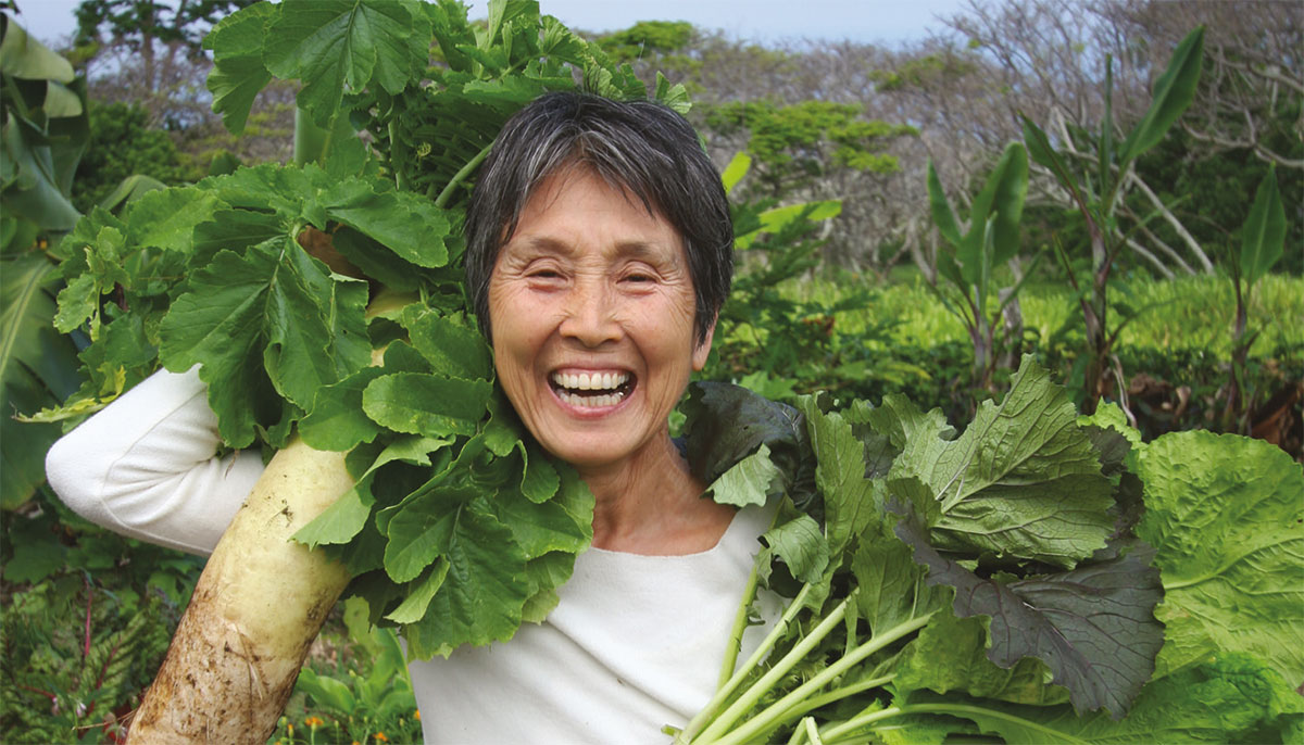 Mayumi Oda smiling in her garden holding up vegetables.
