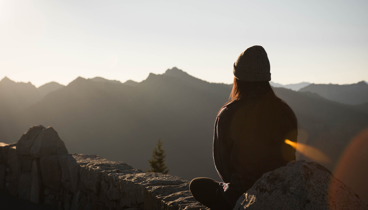 A woman meditating on the edge of a cliff. There are mountains in the background.