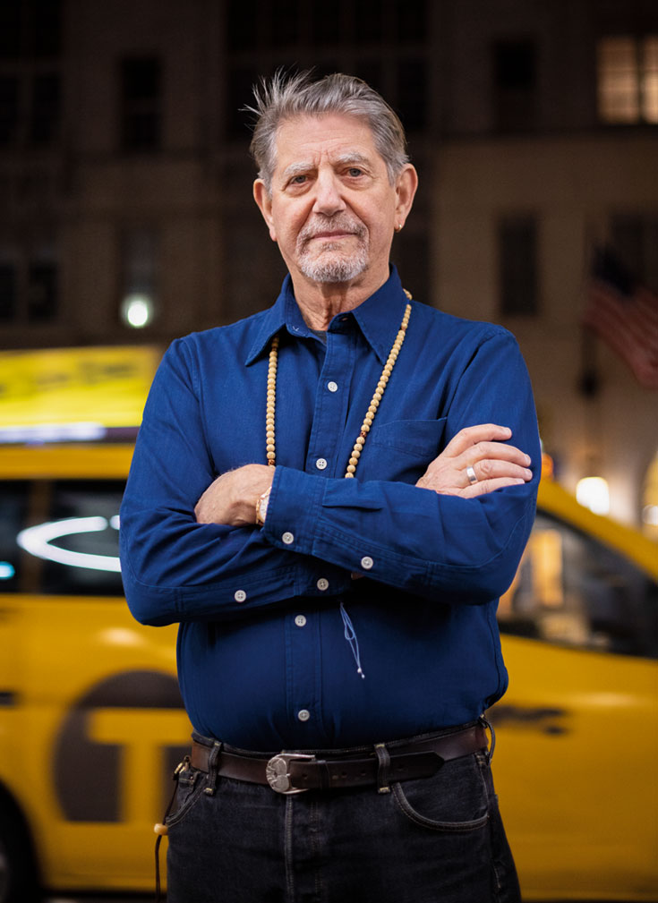 A man standing with his arms crossed in front of a taxi. He is wearing a blue long-sleeve shirt and black pants.