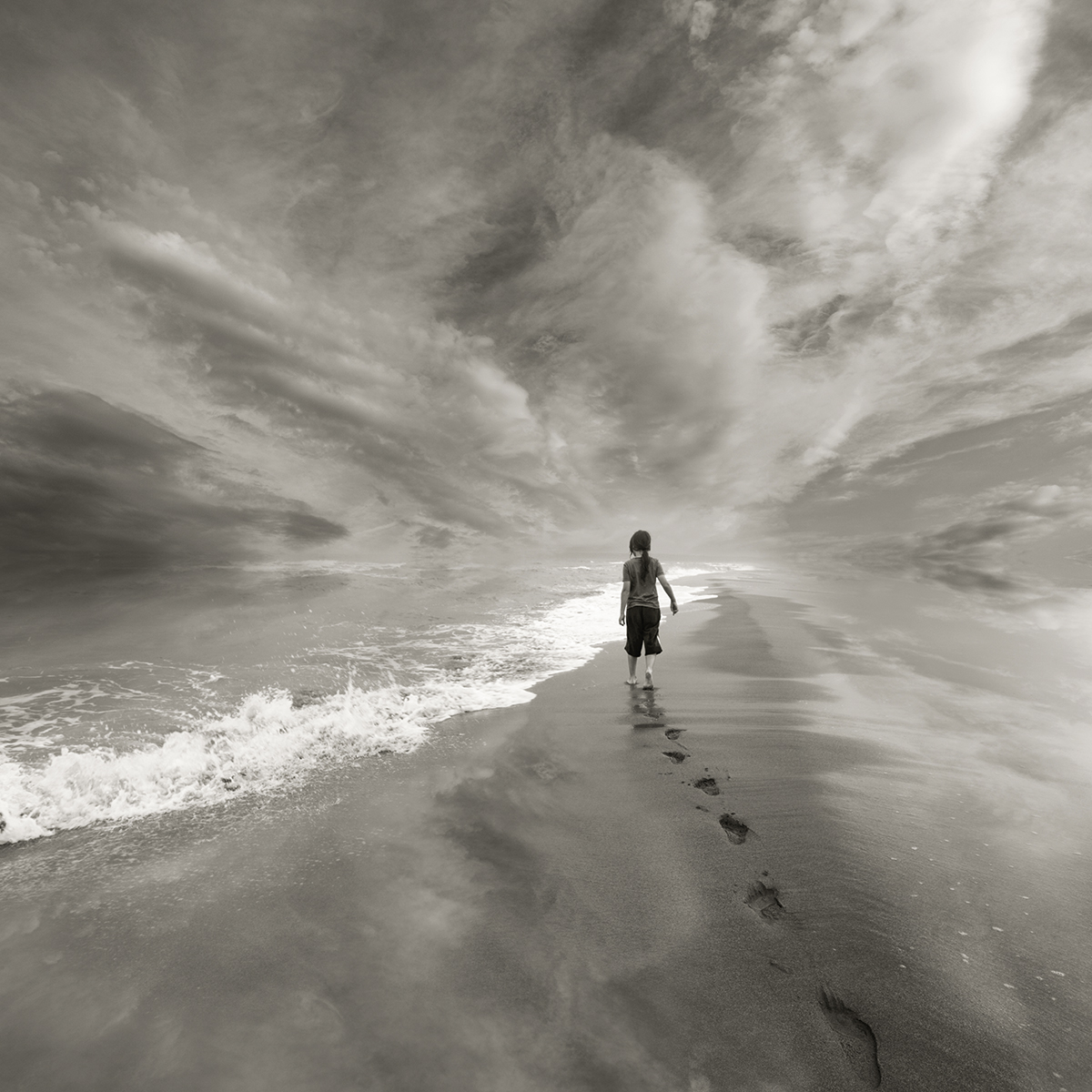 A young person walking down the beach. The photo is in black and white and you can see their footprints.