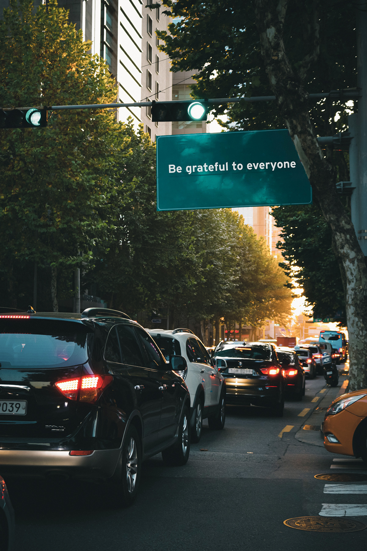 Cars going through a green light. There is a road sign that reads "Be grateful to everyone."