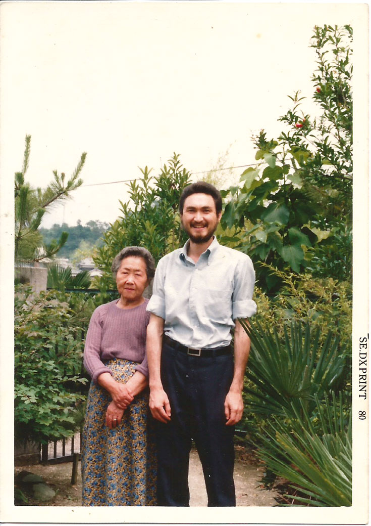 A young man stands next to an older woman in front of plants.