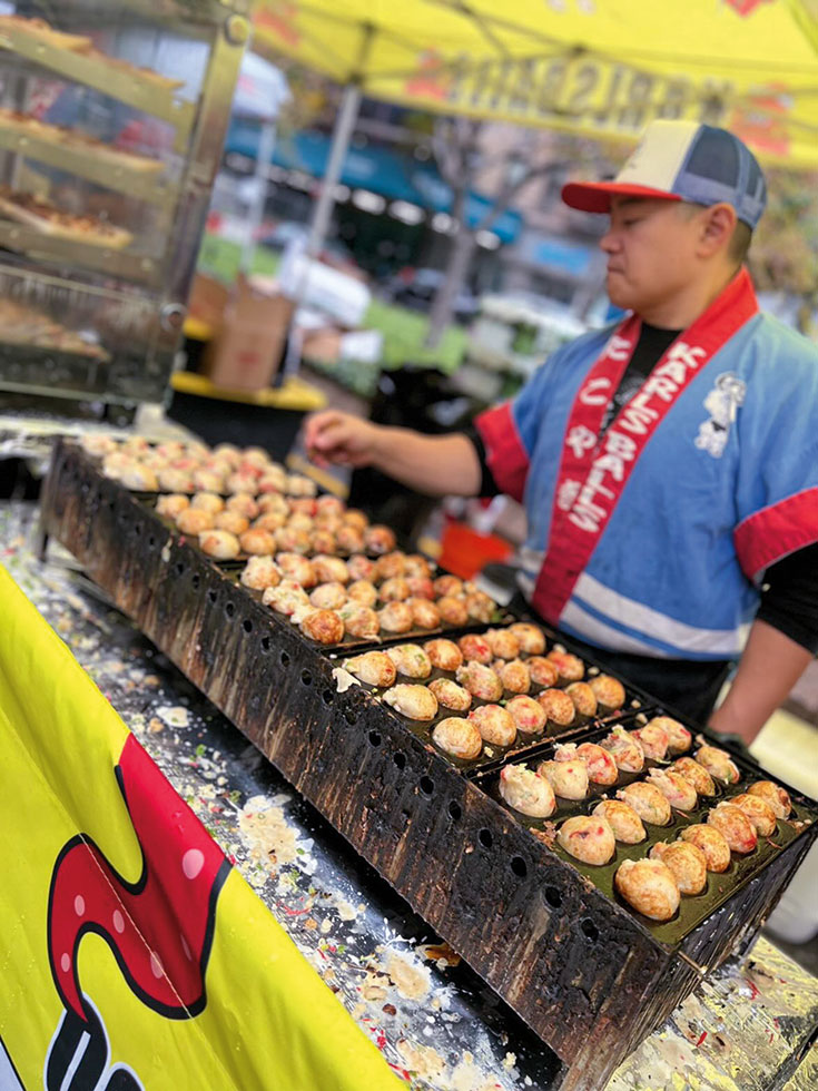Karl Palma wear a blue and red chef's jacket and stands behind his takoyaki stand, turning small balls of dough on a grill.