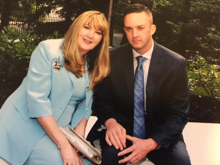 A mother in a blue jacket sits with her son, wearing a suit and tie.