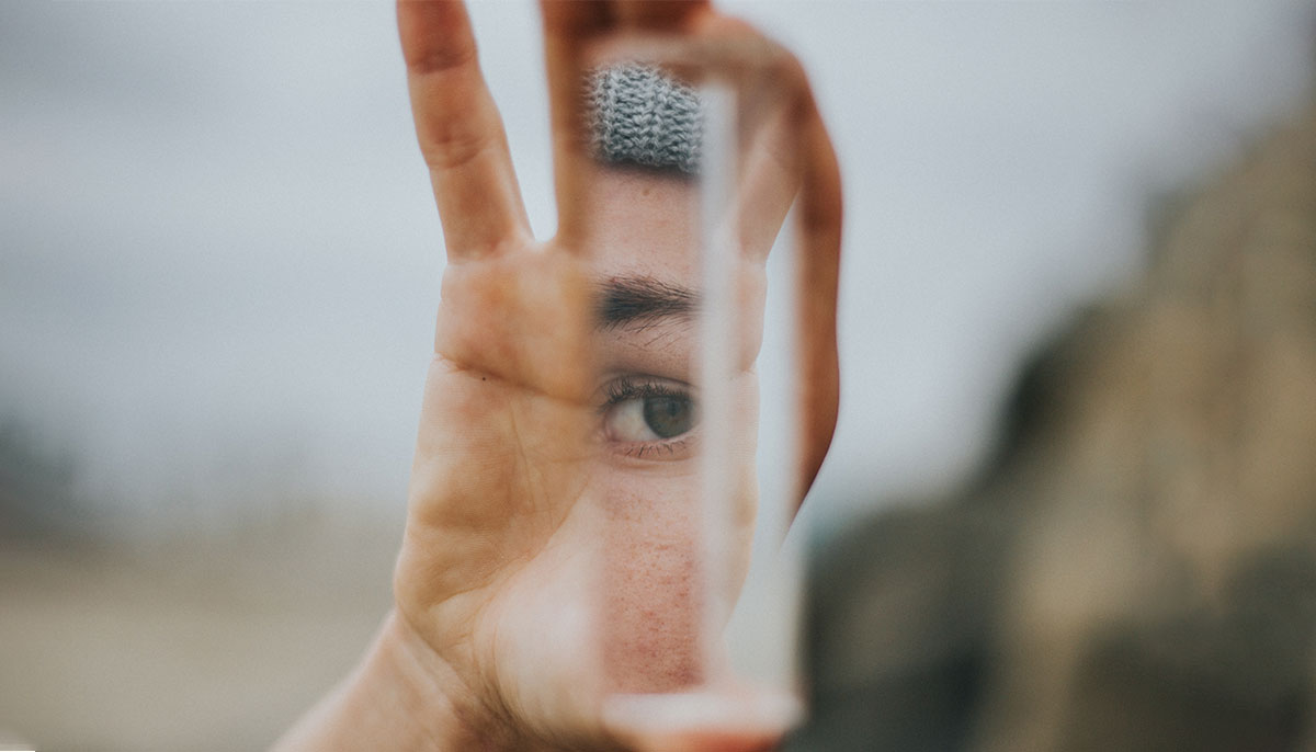 person holds up slice of mirror, looking into their own eye