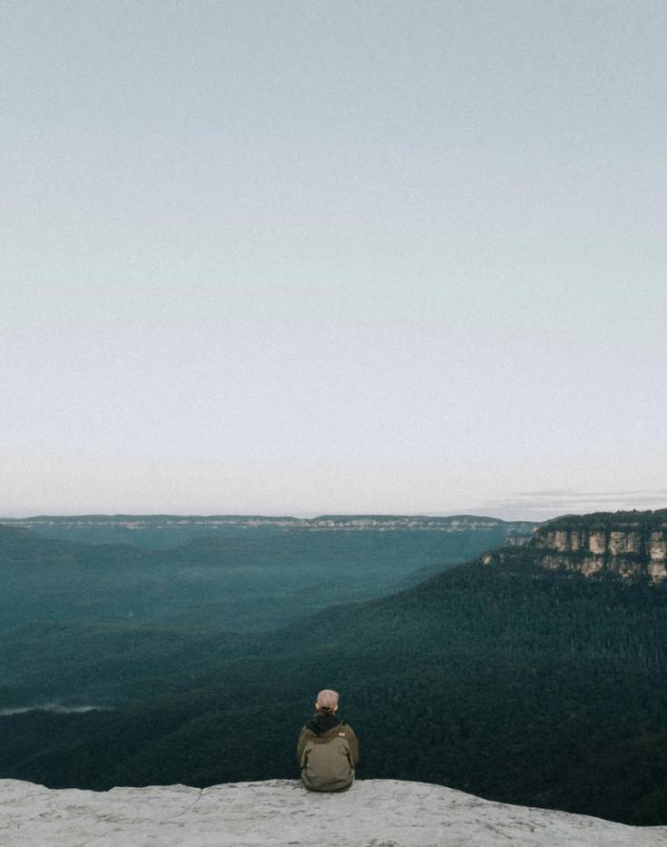 Person sitting on a cliff looking over mountain range.